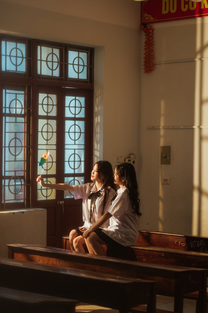 Two young women sitting in a sunlit Vietnamese classroom, wearing uniforms, capturing a photo.